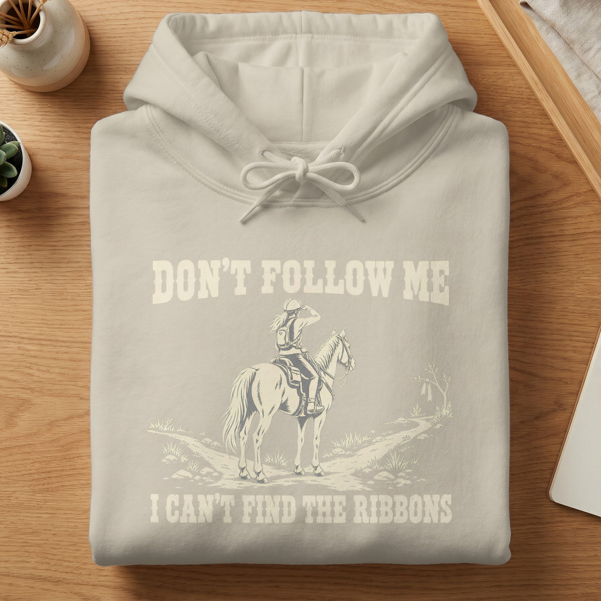 A cream-colored hooded sweatshirt with a graphic of an equestrian rider and the text, Don't Follow Me I Can't Find The Ribbons.