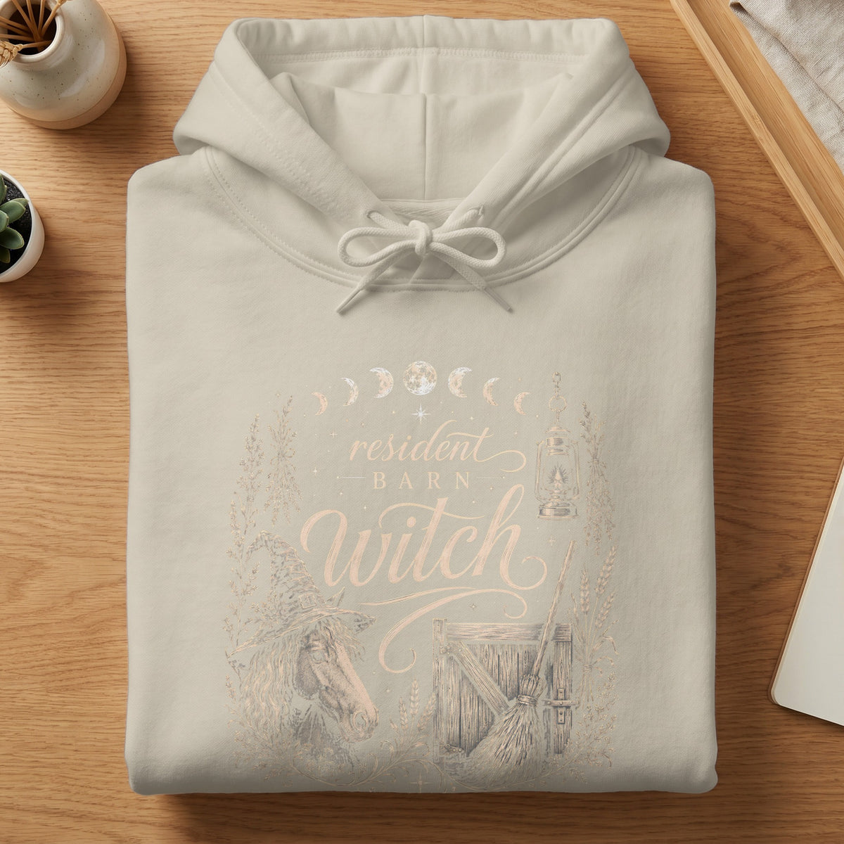 Light tan hoodie with "resident barn witch" graphic featuring horse, broom, and moon phases.