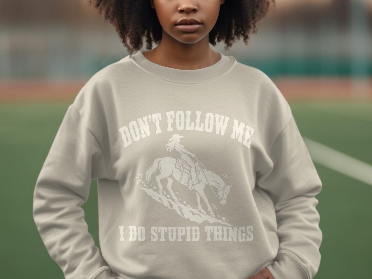 A person wearing a light gray crewneck sweatshirt featuring a graphic of a cowgirl riding a horse with text that reads don't follow me i do stupid things.
