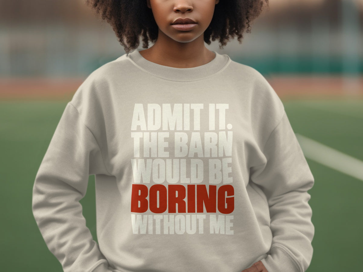 A person wearing a beige crewneck sweatshirt printed with the text ADMIT IT. THE BARN WOULD BE BORING WITHOUT ME.