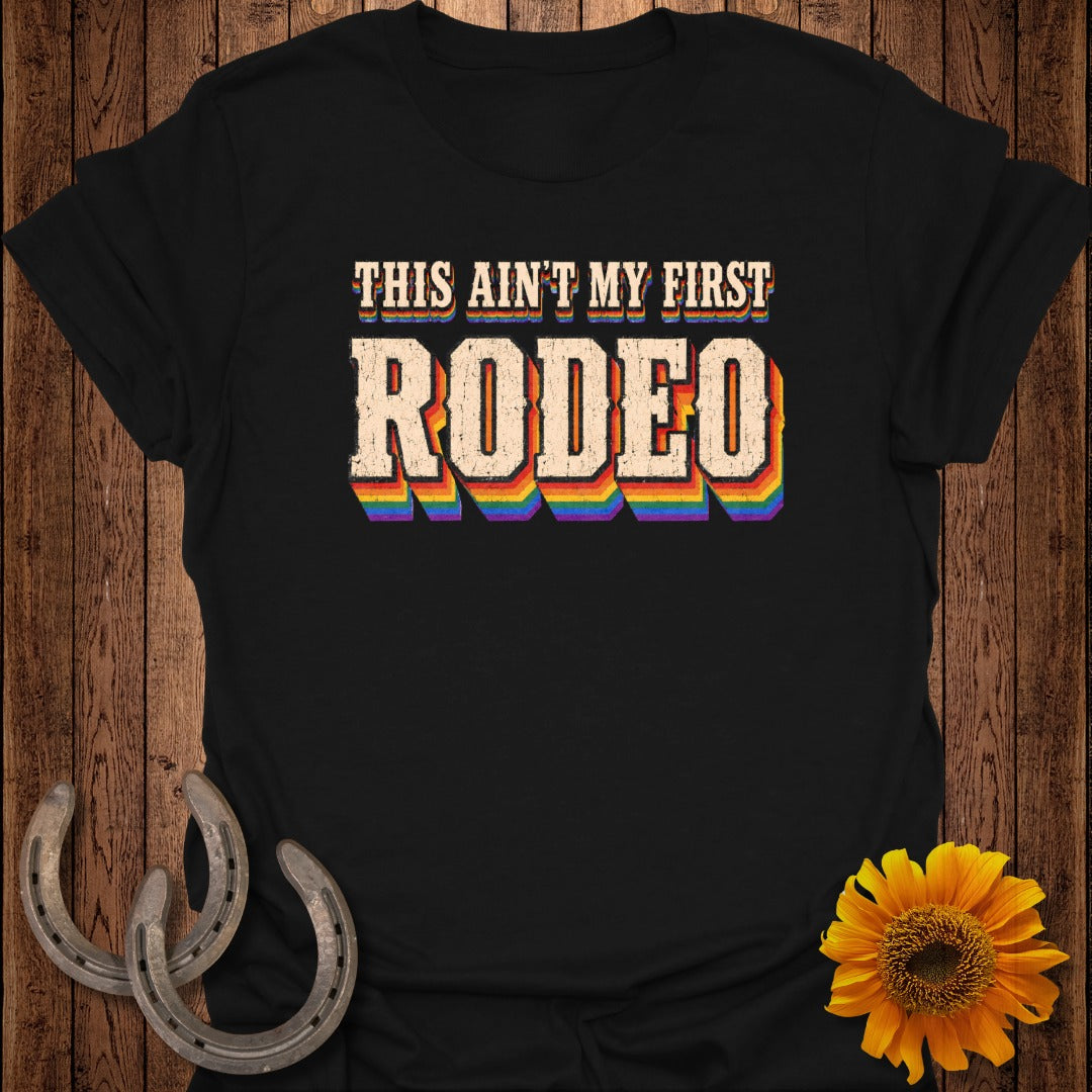 Black t-shirt with "THIS AIN'T MY FIRST RODEO" in rainbow gradient text.