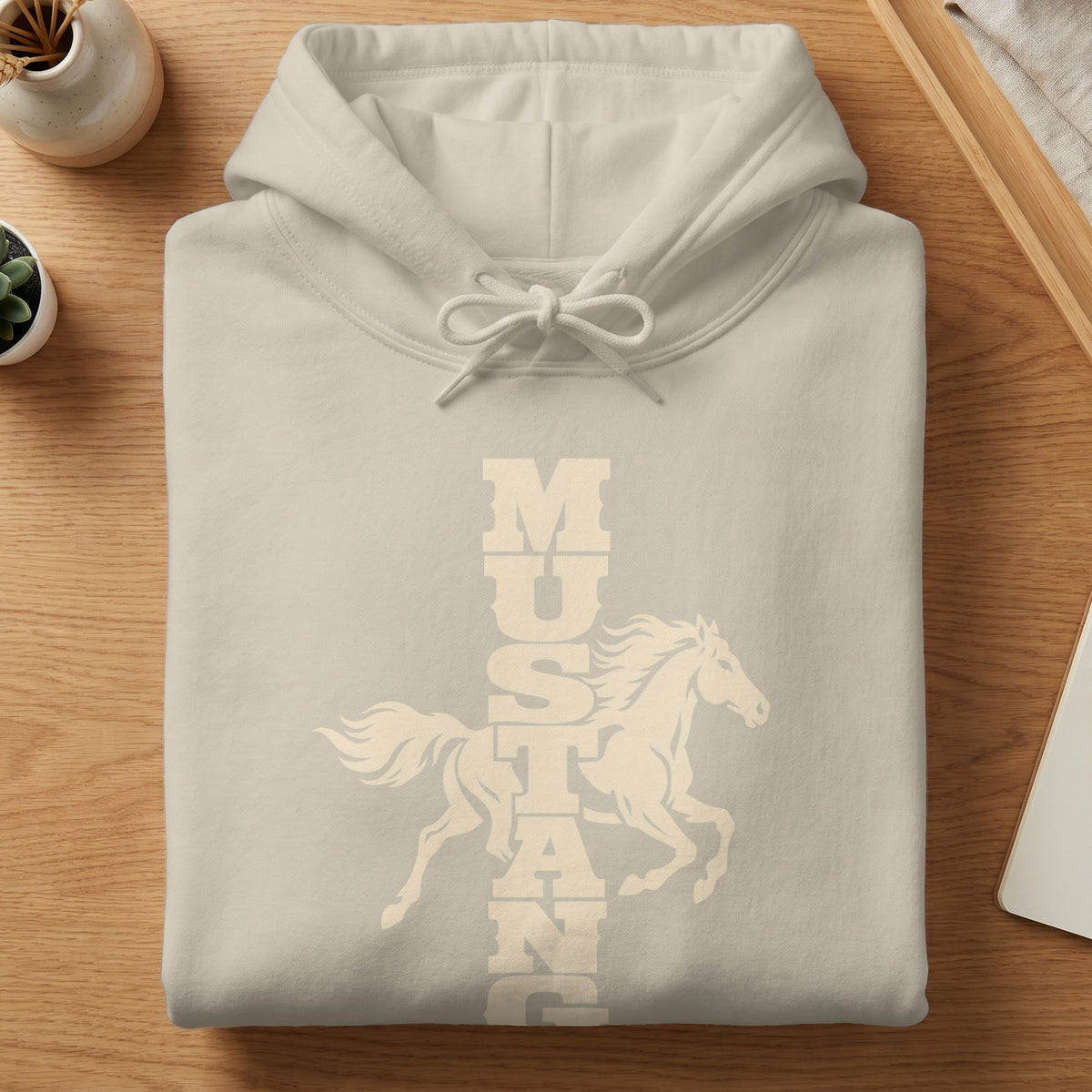 Folded cream-colored hoodie resting on a wooden surface, featuring a graphic of a running horse and vertical text spelling mustang.
